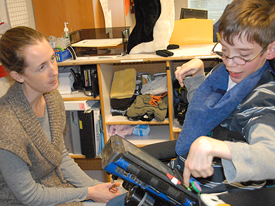 A student in a motorized chair using their AAC device to communicate with a teacher sitting in front of them